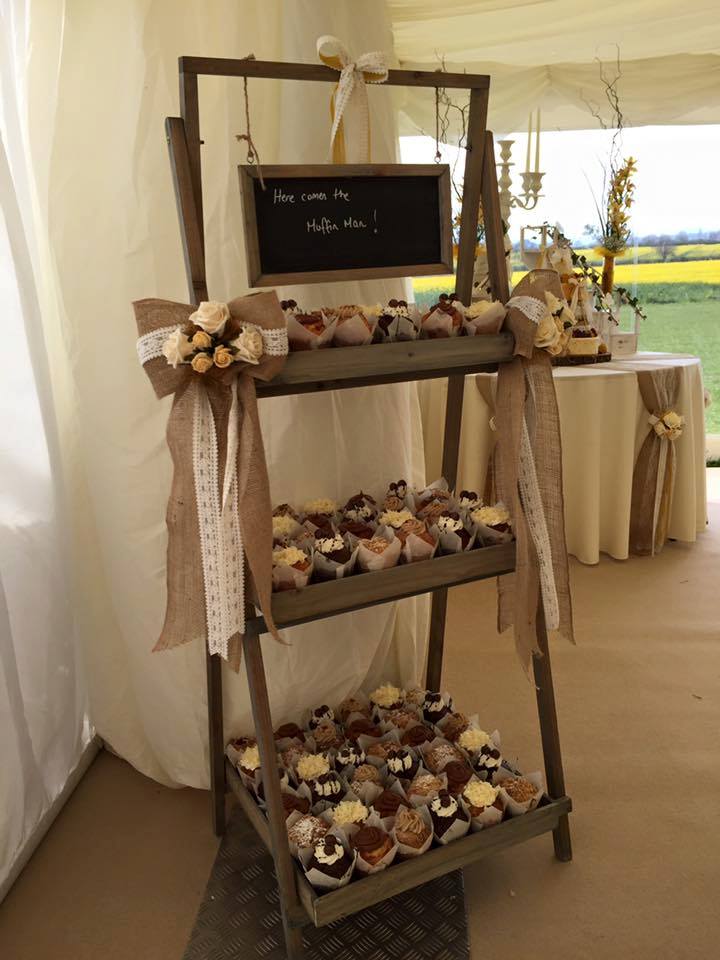 Cupcake display on a wooden ladder shelf with burlap bows, in a tent; yellow field in the background.