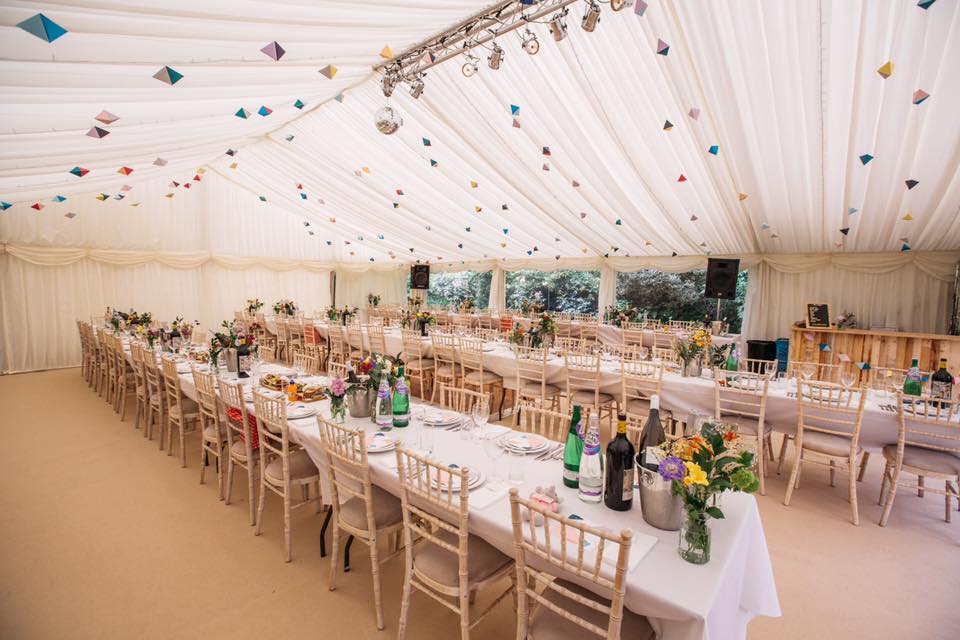 Wedding reception tent with long tables set for dining, floral centerpieces, and decorative hanging squares.