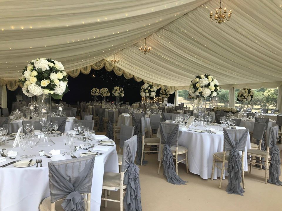 Wedding reception in a tent, tables set with white linens, gray chair sashes, and large floral centerpieces.