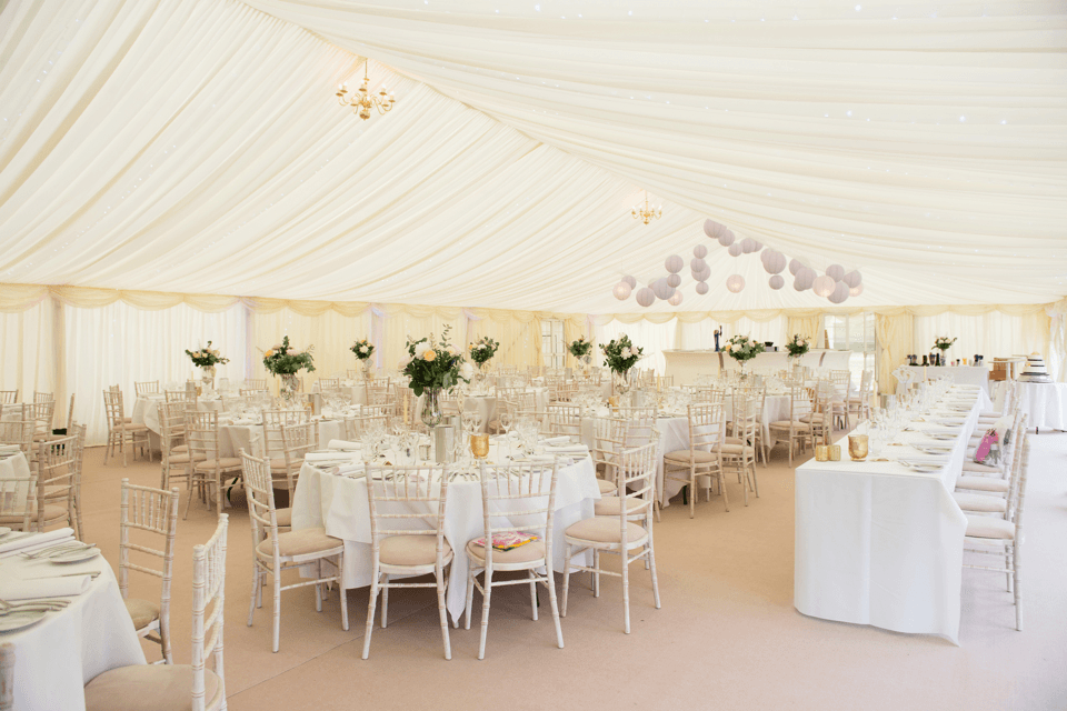 Elegant wedding reception tent with round and rectangular tables, floral centrepieces, and white chairs.