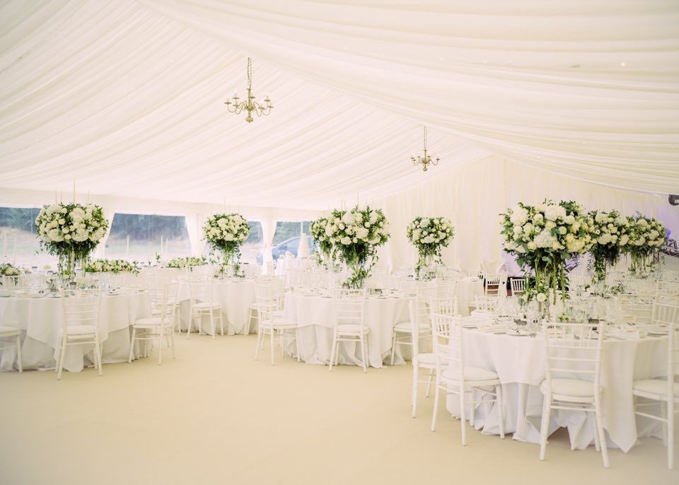 Elegant white tent interior set for a wedding. Round tables with floral centerpieces, white chairs, and chandeliers.