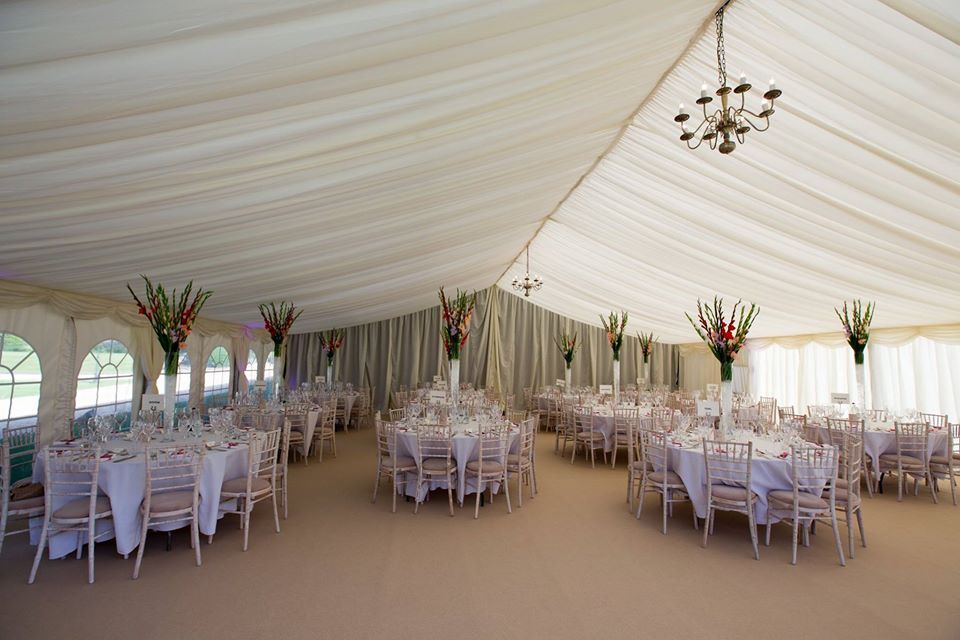 Interior of a wedding tent. Round tables with white linens, flowers, and chairs are arranged for a reception.