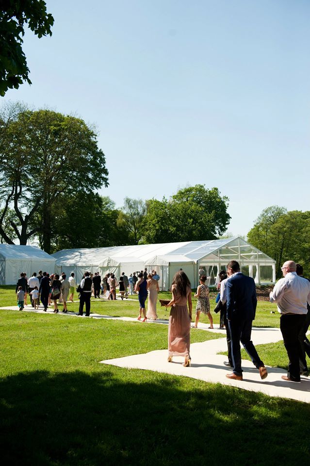 People walking on a grassy lawn toward a white tent on a sunny day.