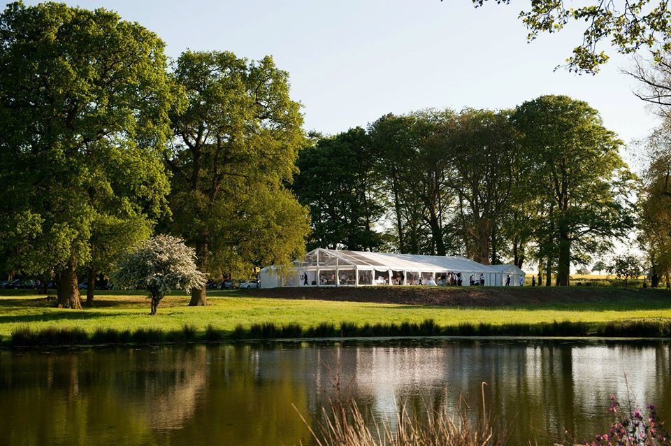 White tent on a grassy bank by a lake, trees in the background, sunny day.