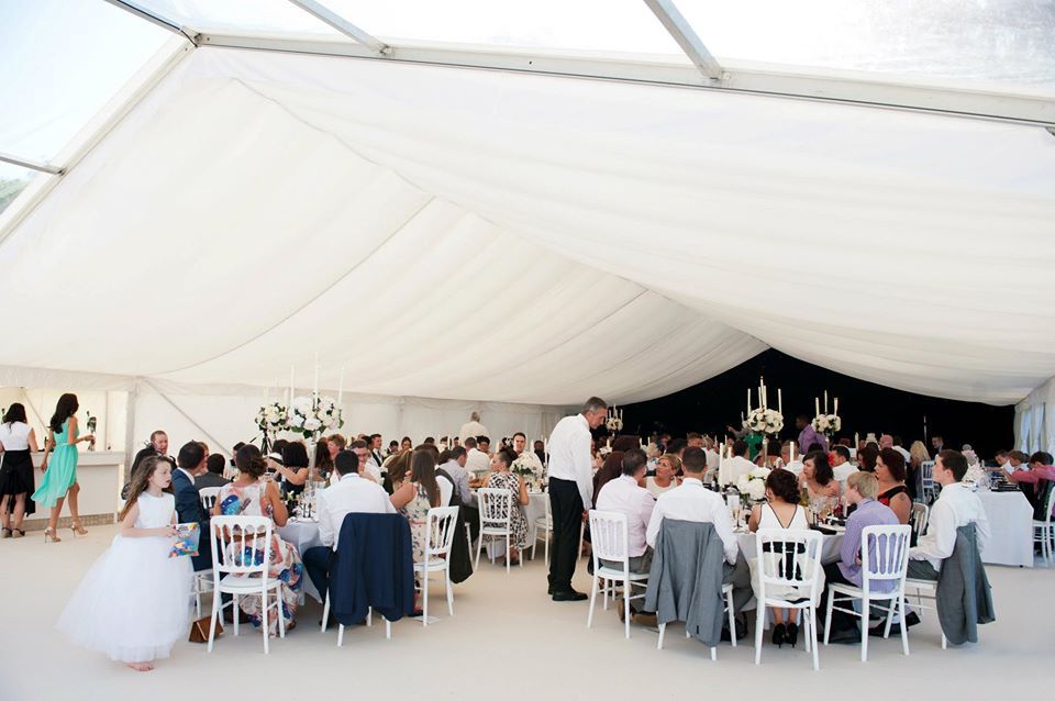 Wedding reception in a white tent, guests seated at tables, a flower girl walks past.