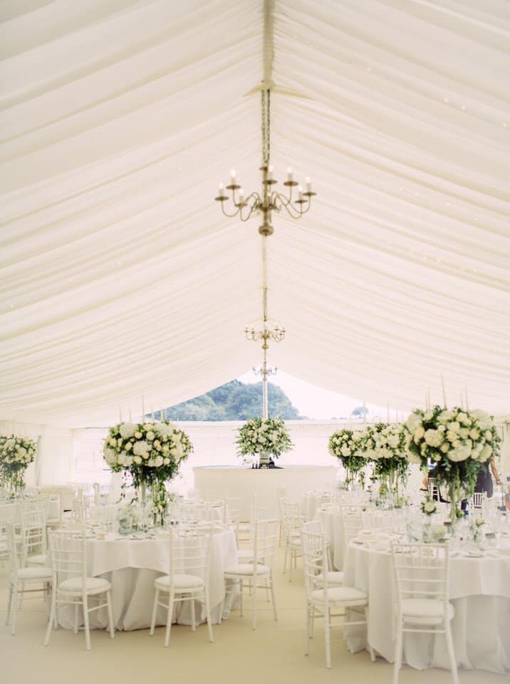 Elegant white wedding reception tent with round tables, floral centerpieces, and a chandelier.