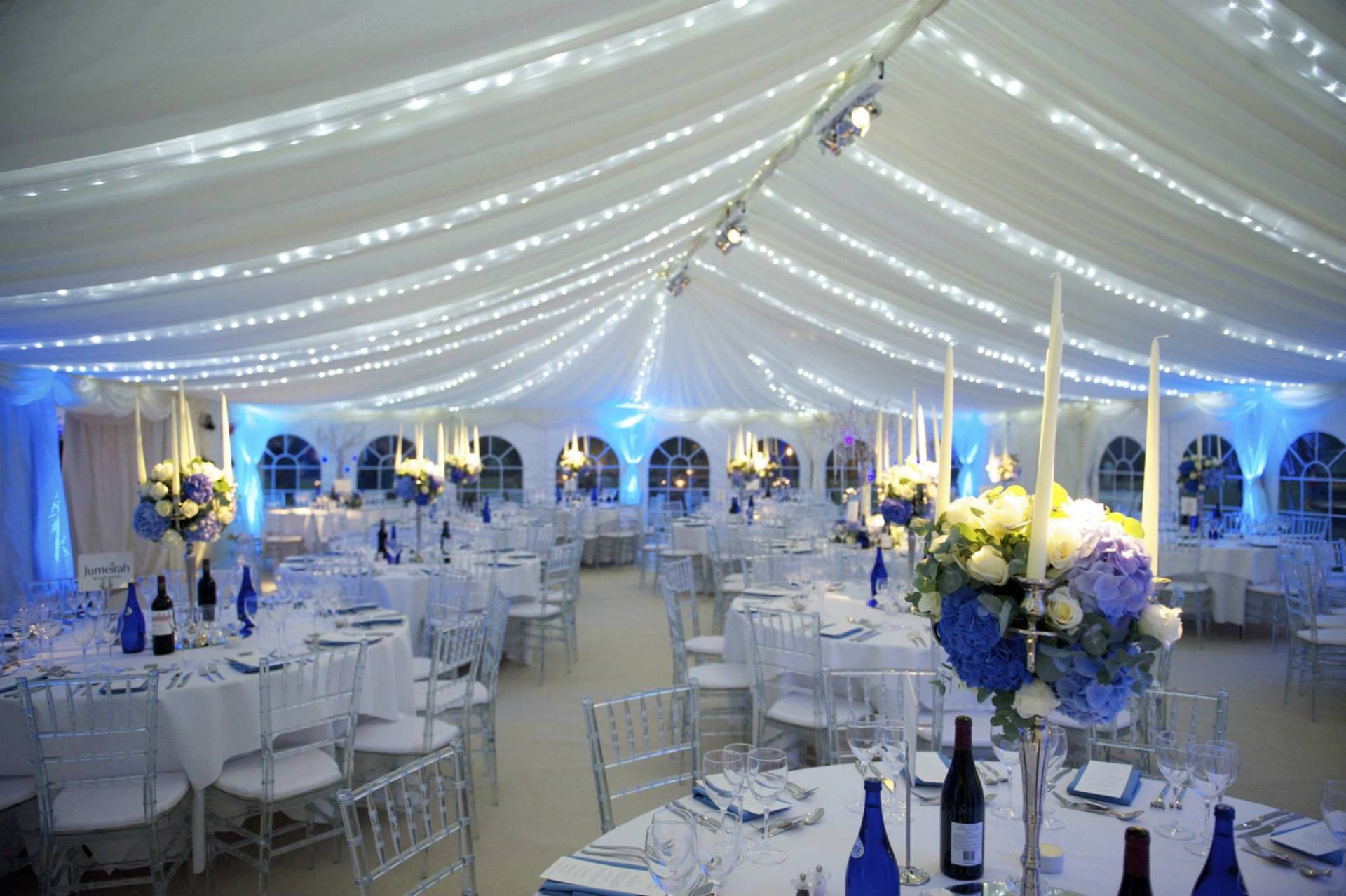 Elegant tent interior decorated for an event with white and blue floral centerpieces and tables set for dining.