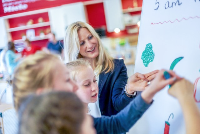 Eine Frau bringt einer Gruppe von Kindern bei, wie man auf einer weißen Tafel zeichnet.