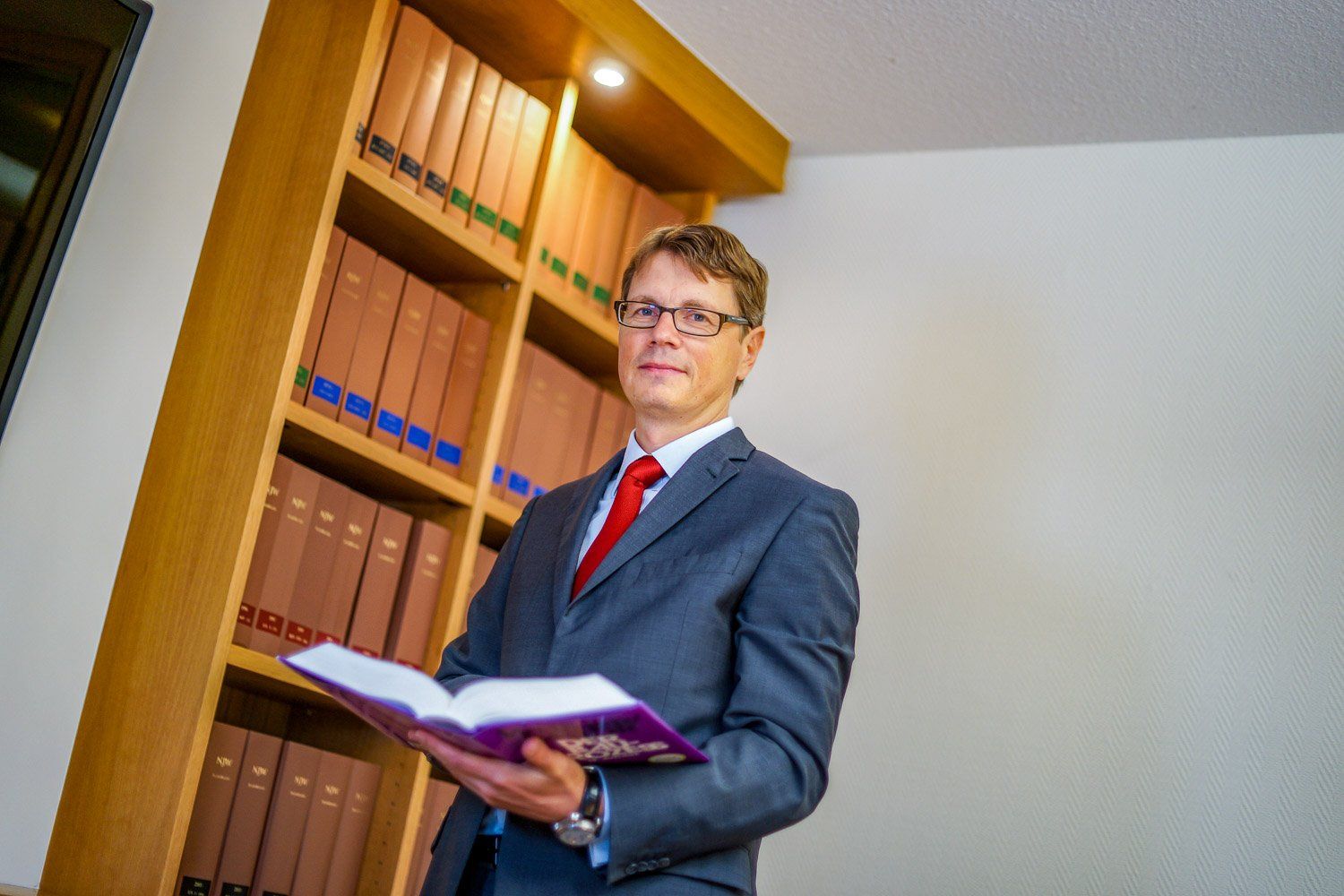 Ein Mann in Anzug und Krawatte hält ein Buch vor einem Bücherregal.
