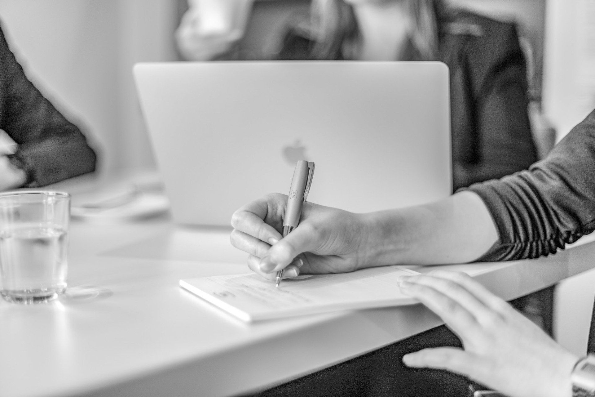 Eine Person sitzt mit einem Laptop an einem Tisch und schreibt auf einem Blatt Papier.
