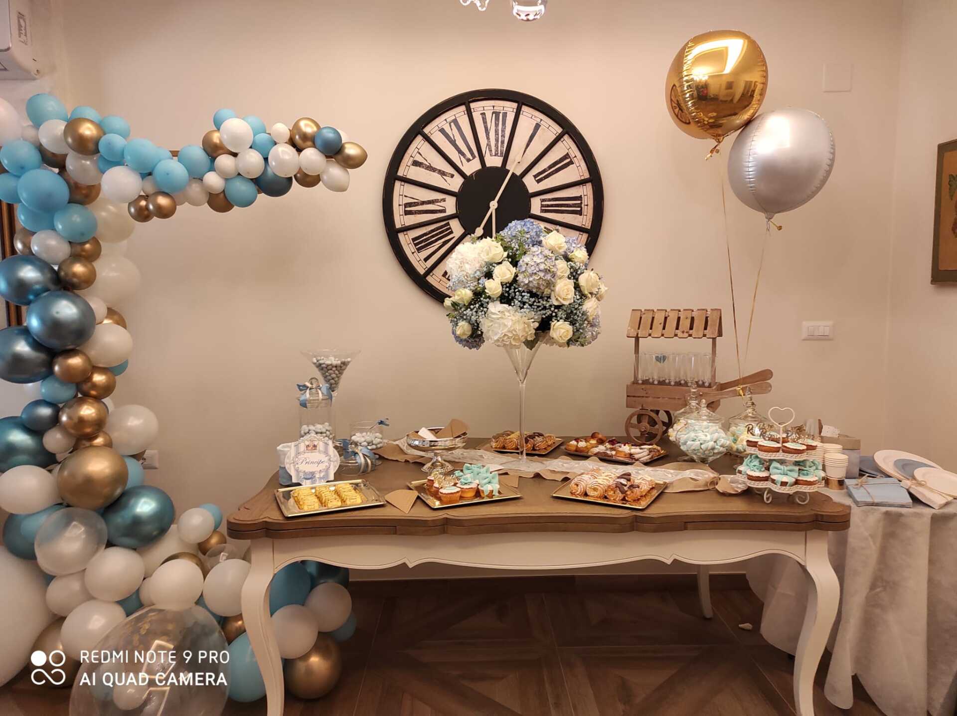 Tavolo per feste con palloncini, orologio e dolci. Colori blu, oro e bianco.