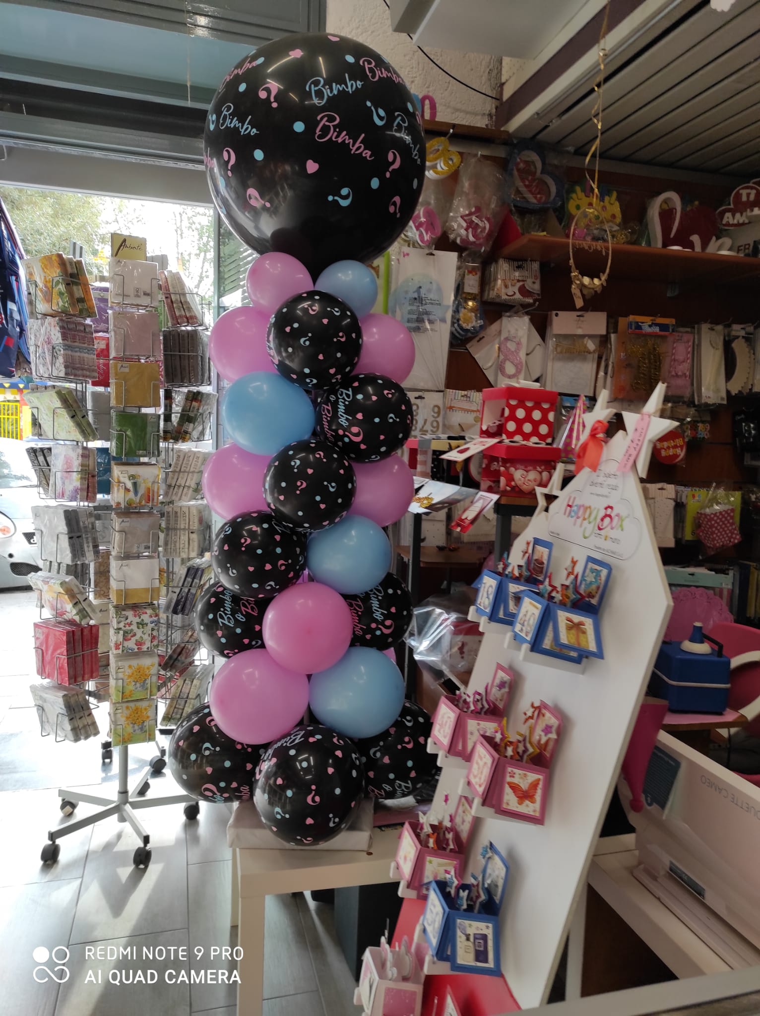 Una colonna di palloncini rosa, blu e neri, sormontata da un grande palloncino floreale, esposta all'interno di un negozio.