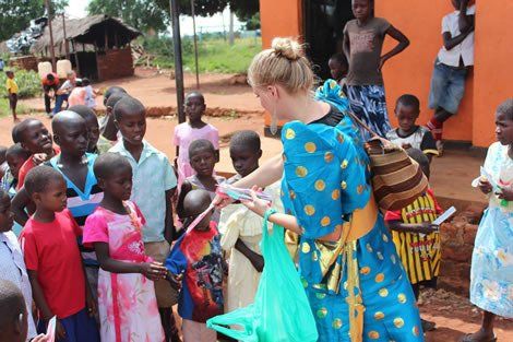 Woman in blue dress giving items to a group of African children, outdoors.