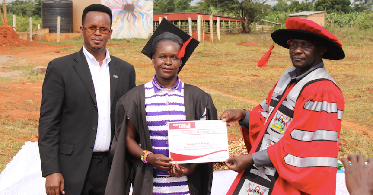 A woman in graduation attire receives a certificate from a person in academic robes, another man stands by. Outdoors.