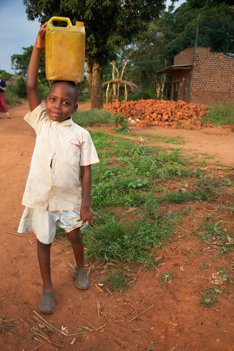 Boy with a yellow water jug balanced on his head in a rural setting.