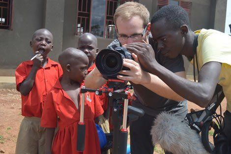 Filmmakers in Uganda filming children in red uniforms. Man looks through the camera with another man.