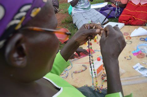 Woman stringing beads, crafting jewelry outdoors, hands focused, wearing glasses and a green top.
