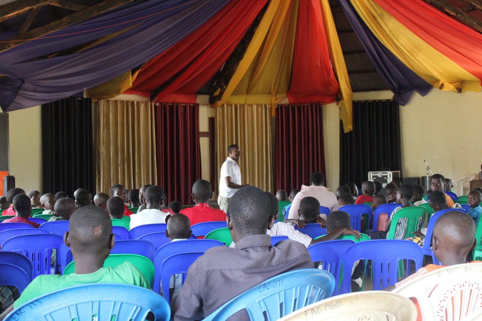 A man speaks to a seated audience in a hall decorated with fabric drapes.