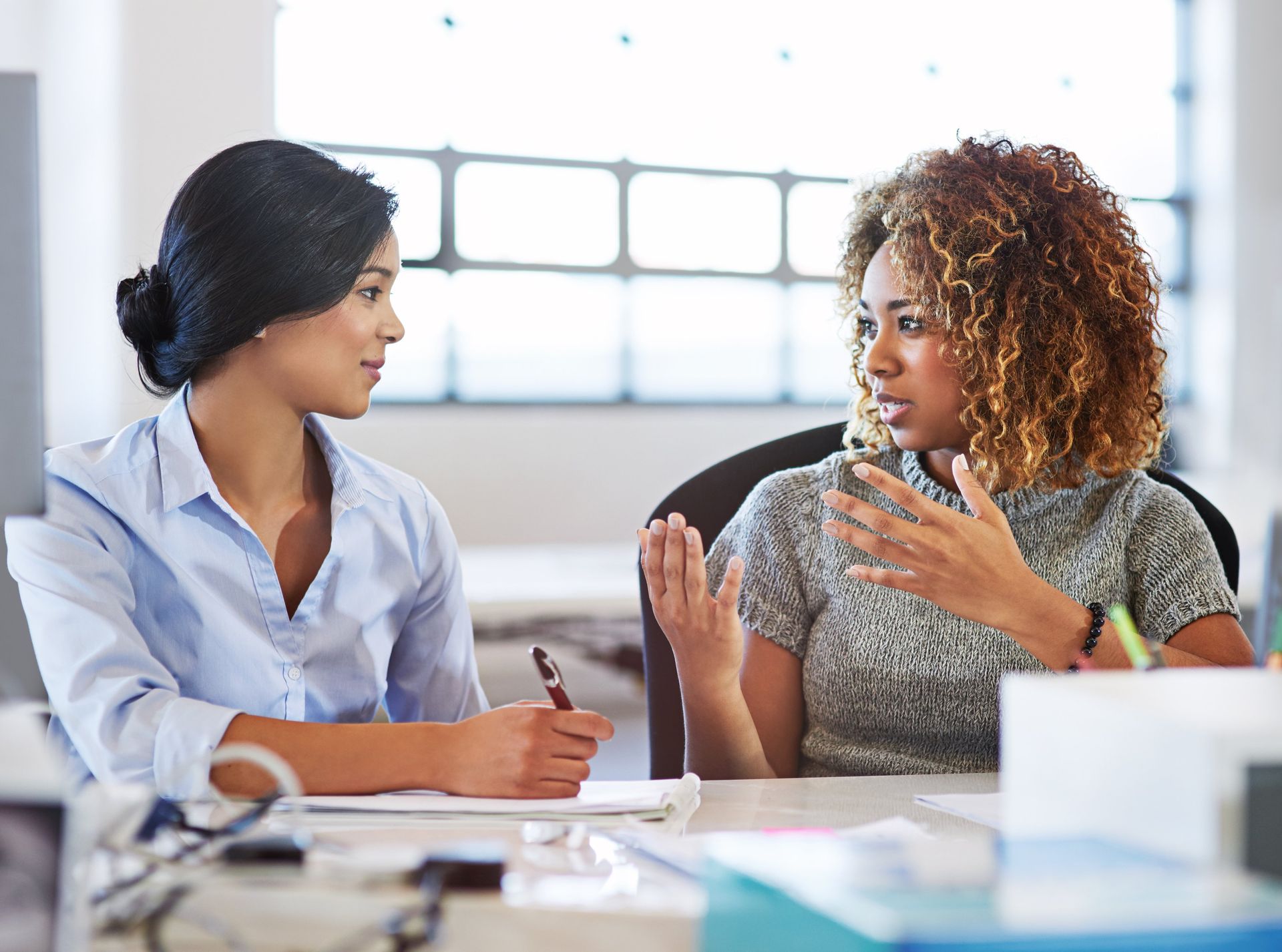 Two women are sitting at a desk talking to each other.