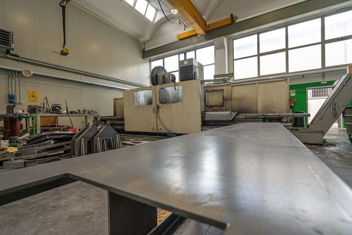Ampio tavolo metallico in un'officina industriale con macchinari, finestre e luci di una gru a ponte.