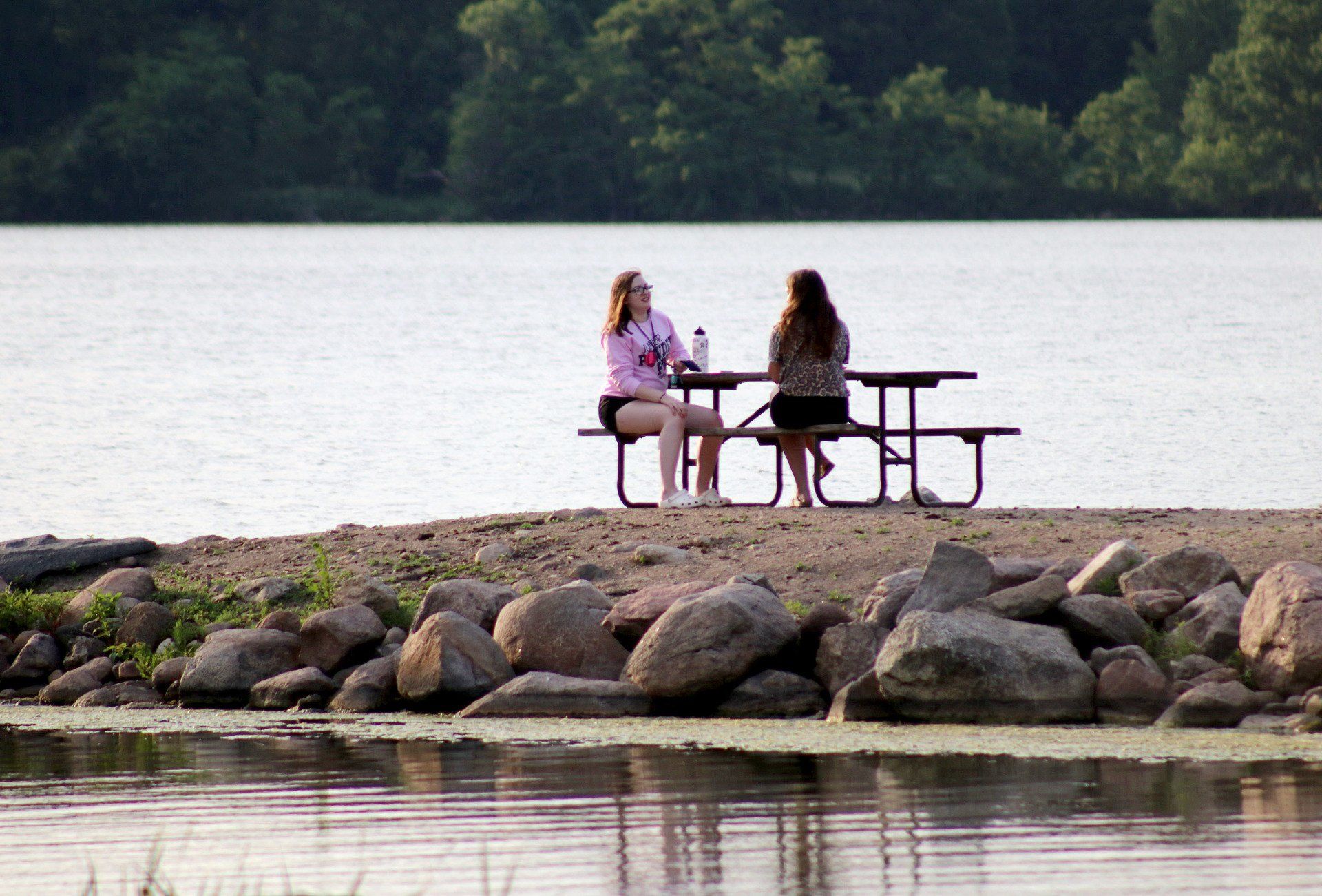Two women are sitting at a picnic table on the shore of a lake.