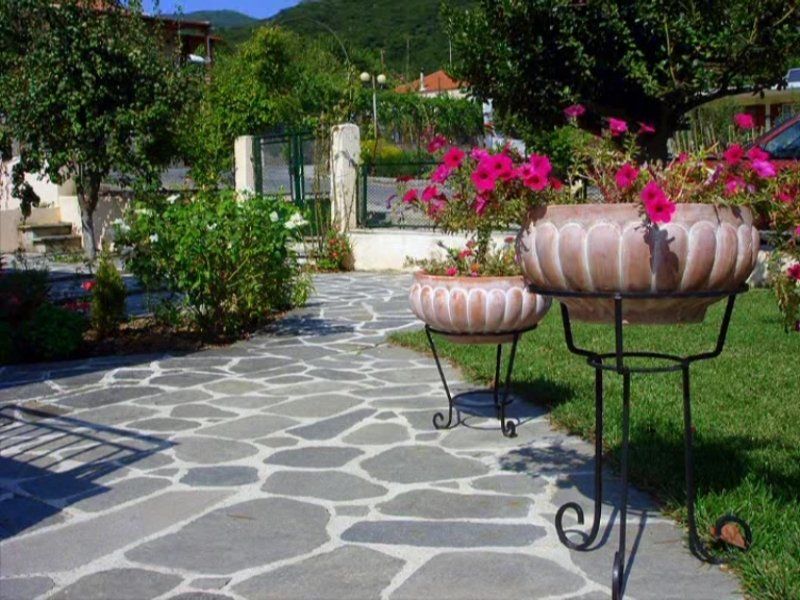 Stone walkway with flower pots on stands, in a garden with green grass and trees.