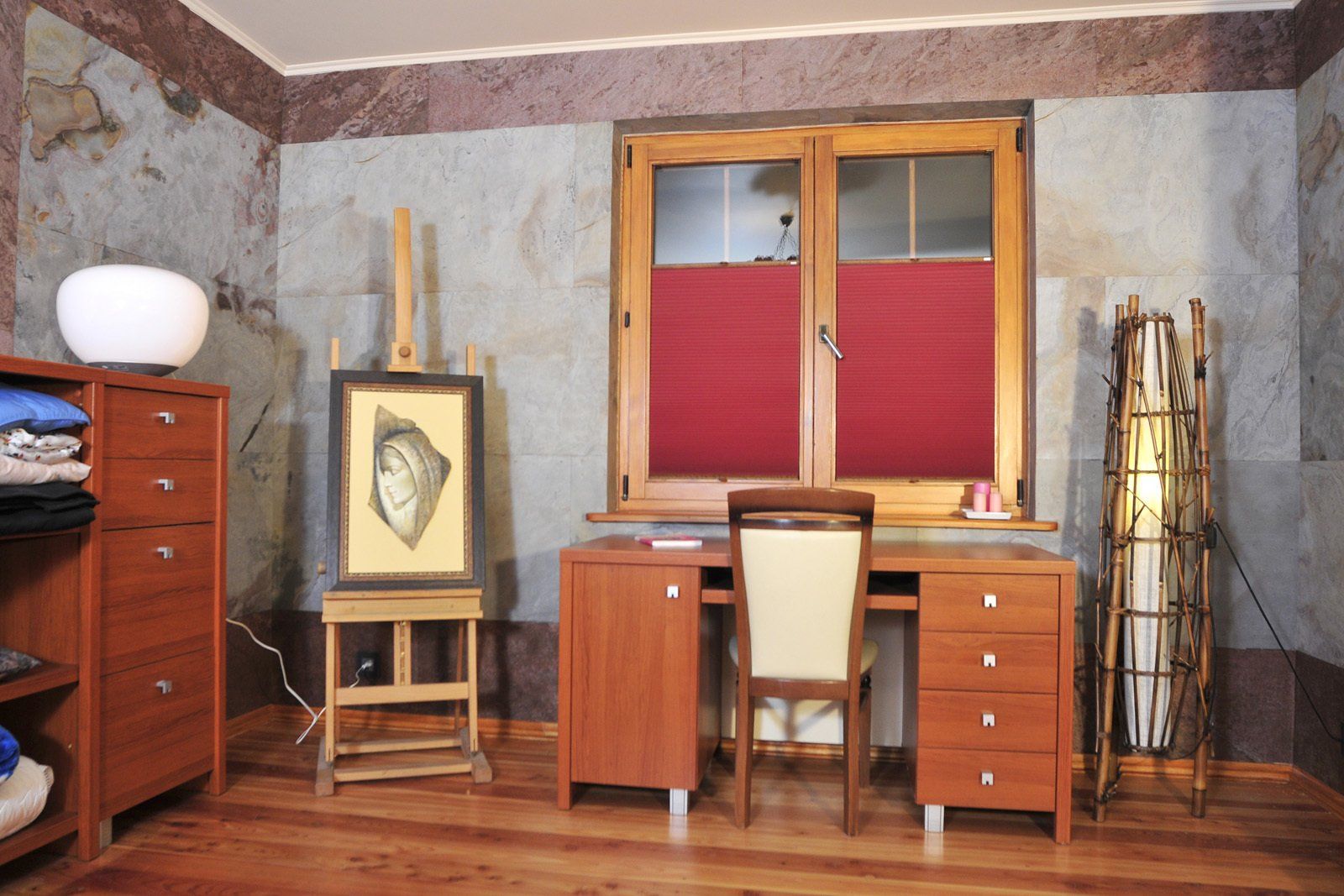 Home office with a desk, window, painting on an easel, lamp, and cabinetry against stone walls.