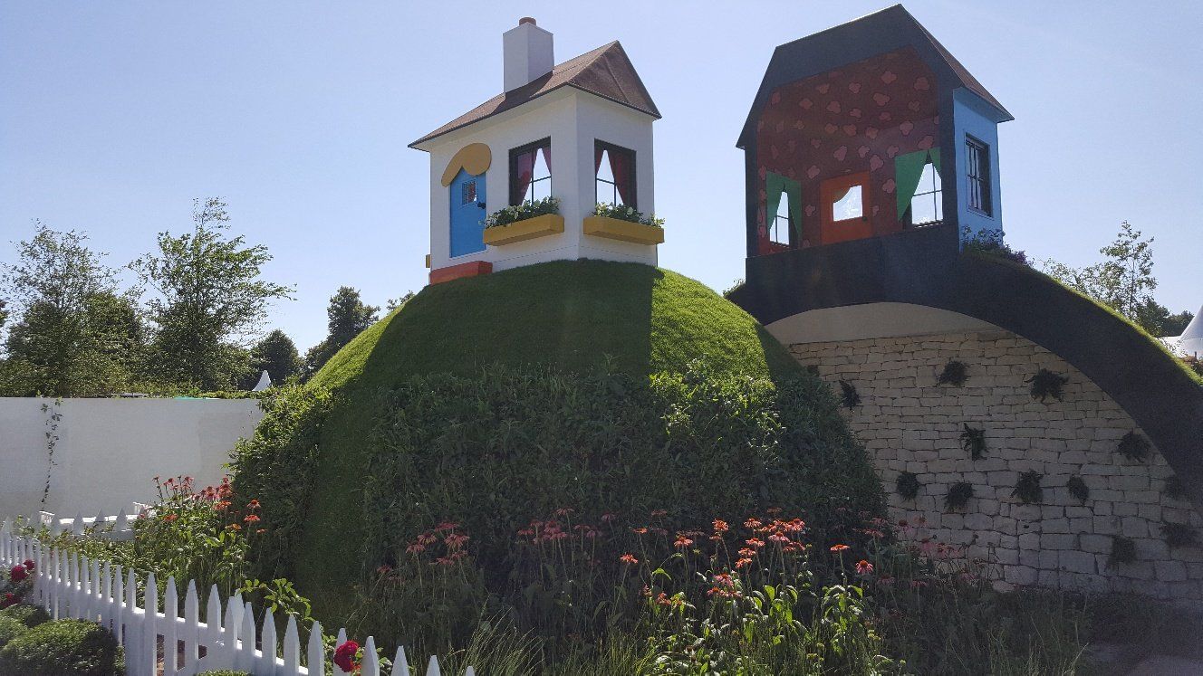 Miniature storybook houses atop a grassy hill with a white picket fence under a blue sky.