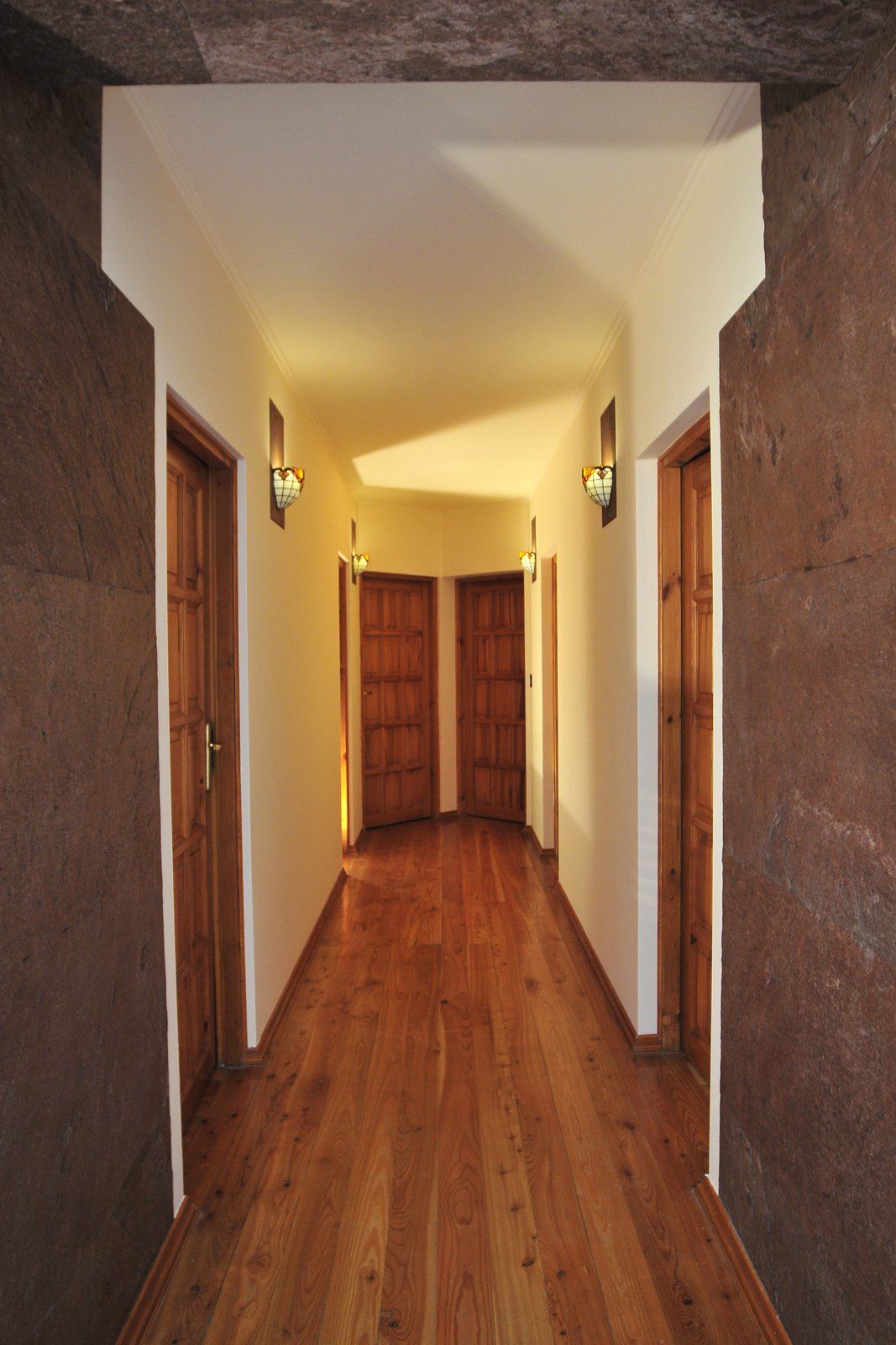 Hallway with wooden floor, doors, and sconce lighting; stone walls on the sides and white ceiling.