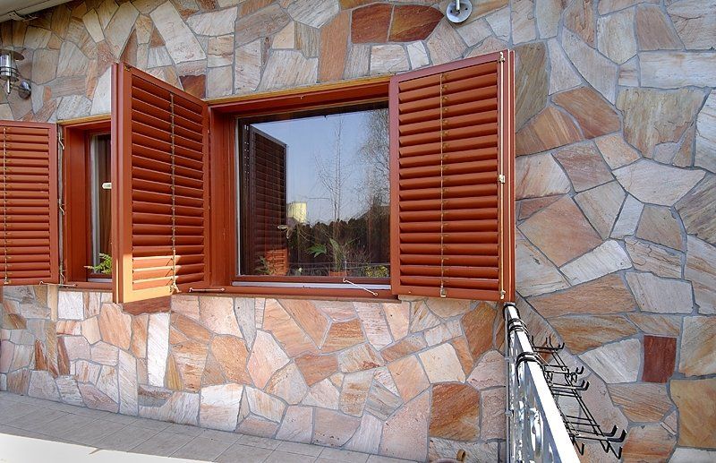 Window with brown shutters on a stone-covered exterior wall; shutters are open.