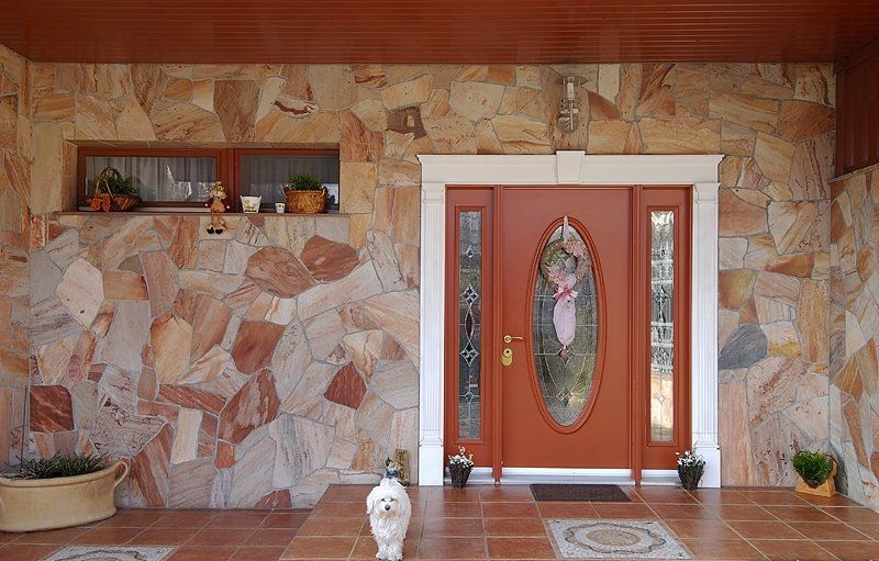 Brown front door with oval glass and flanking side panels, on a stone-clad exterior with a small dog in front.