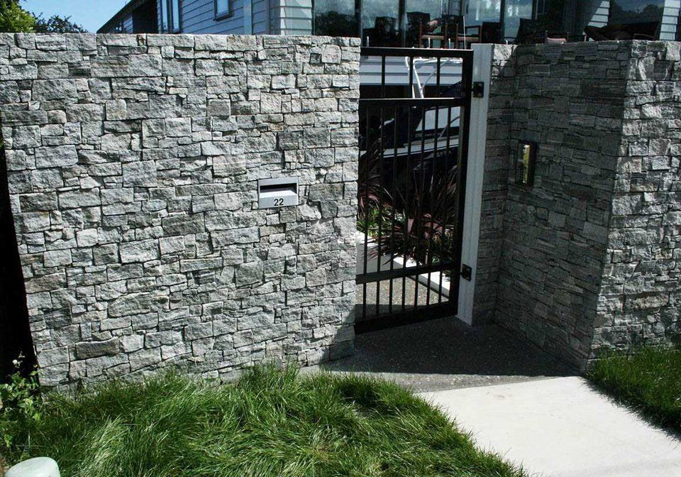 Stone wall with black iron gate leading to a house, with green grass in foreground.