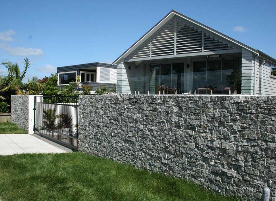 Gray stone wall in front of a modern house with a sloped roof and glass doors, on a sunny day.