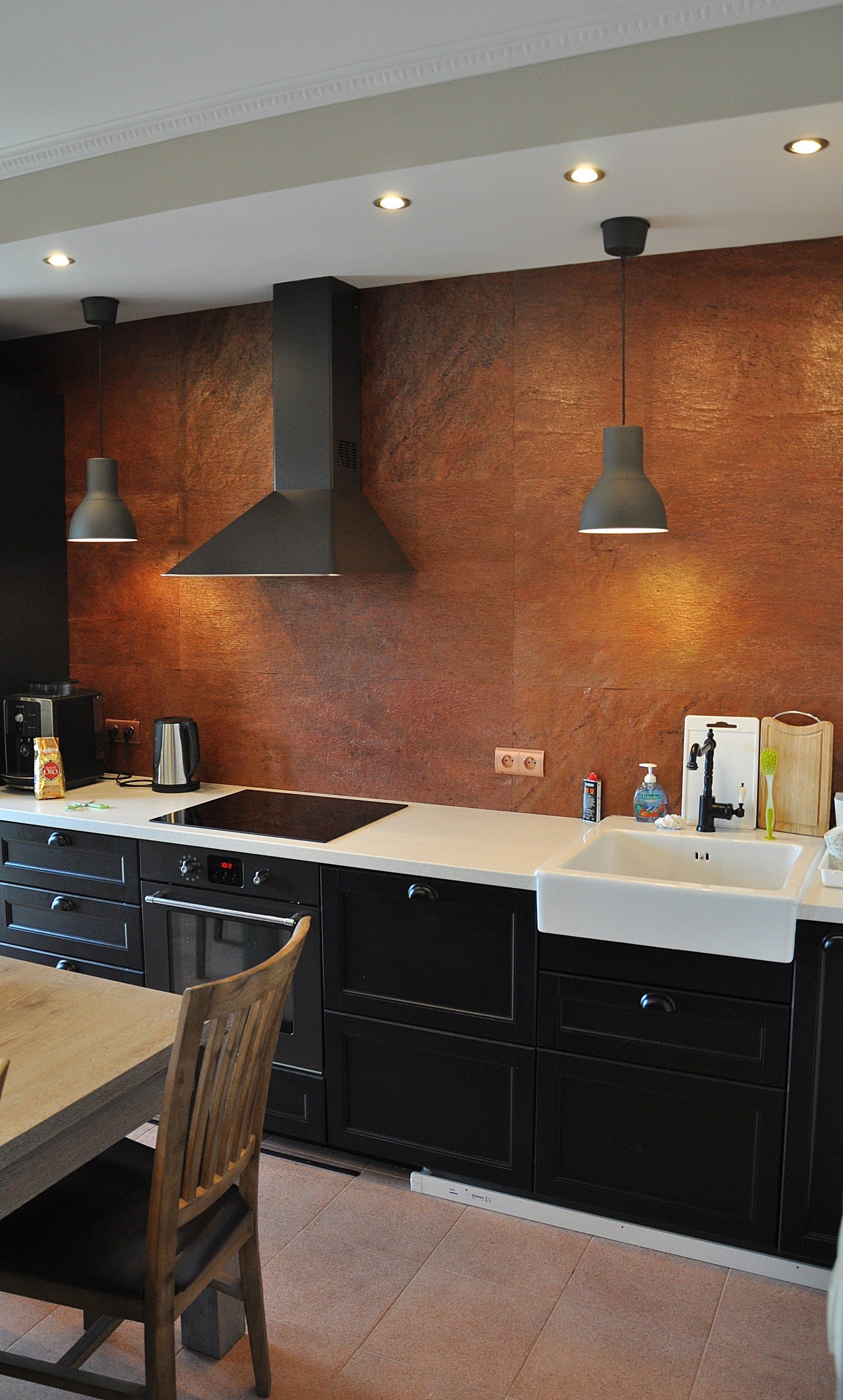 Kitchen with copper backsplash, black cabinets, white sink, and gray pendant lights.