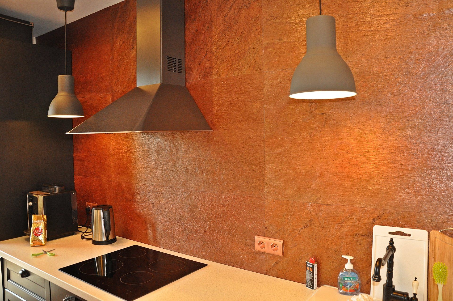 Kitchen with copper-toned backsplash, black range hood, stovetop, and two pendant lights.