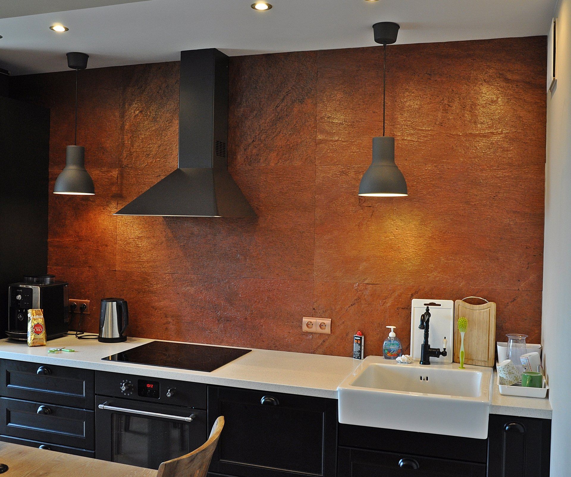 Kitchen with black cabinets, white countertop, copper-toned backsplash, black range hood, and pendant lights.