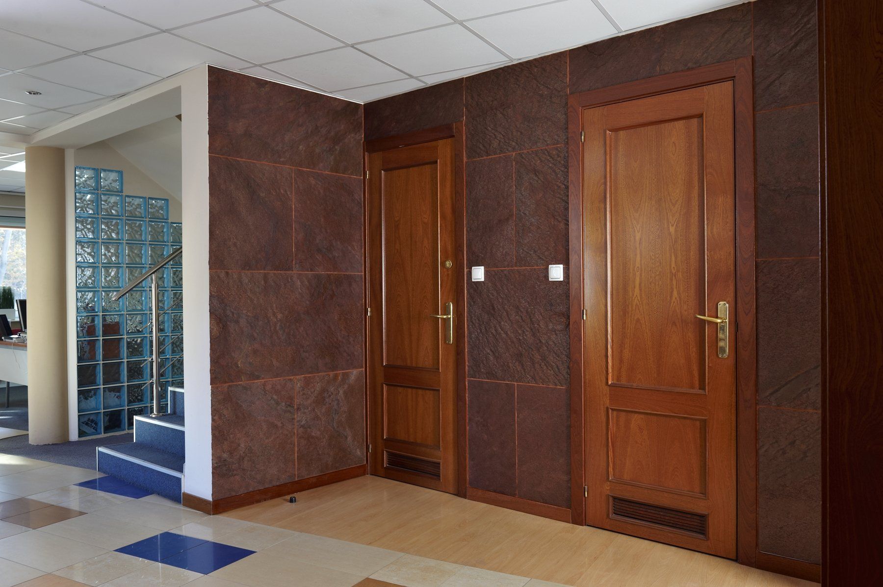 Interior view of a hallway with two wooden doors, brown textured walls, and a staircase with glass block accents.