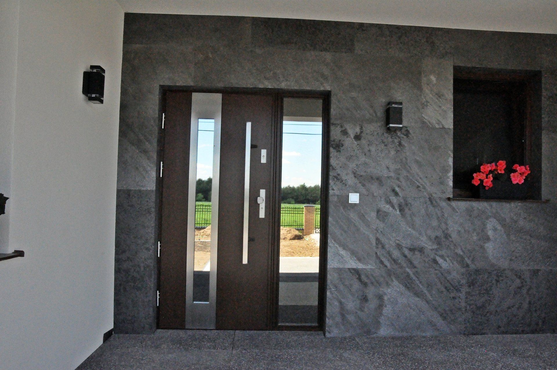 Front door with dark wood and stainless steel accents, flanked by textured gray stone. Red flowers in a small window.