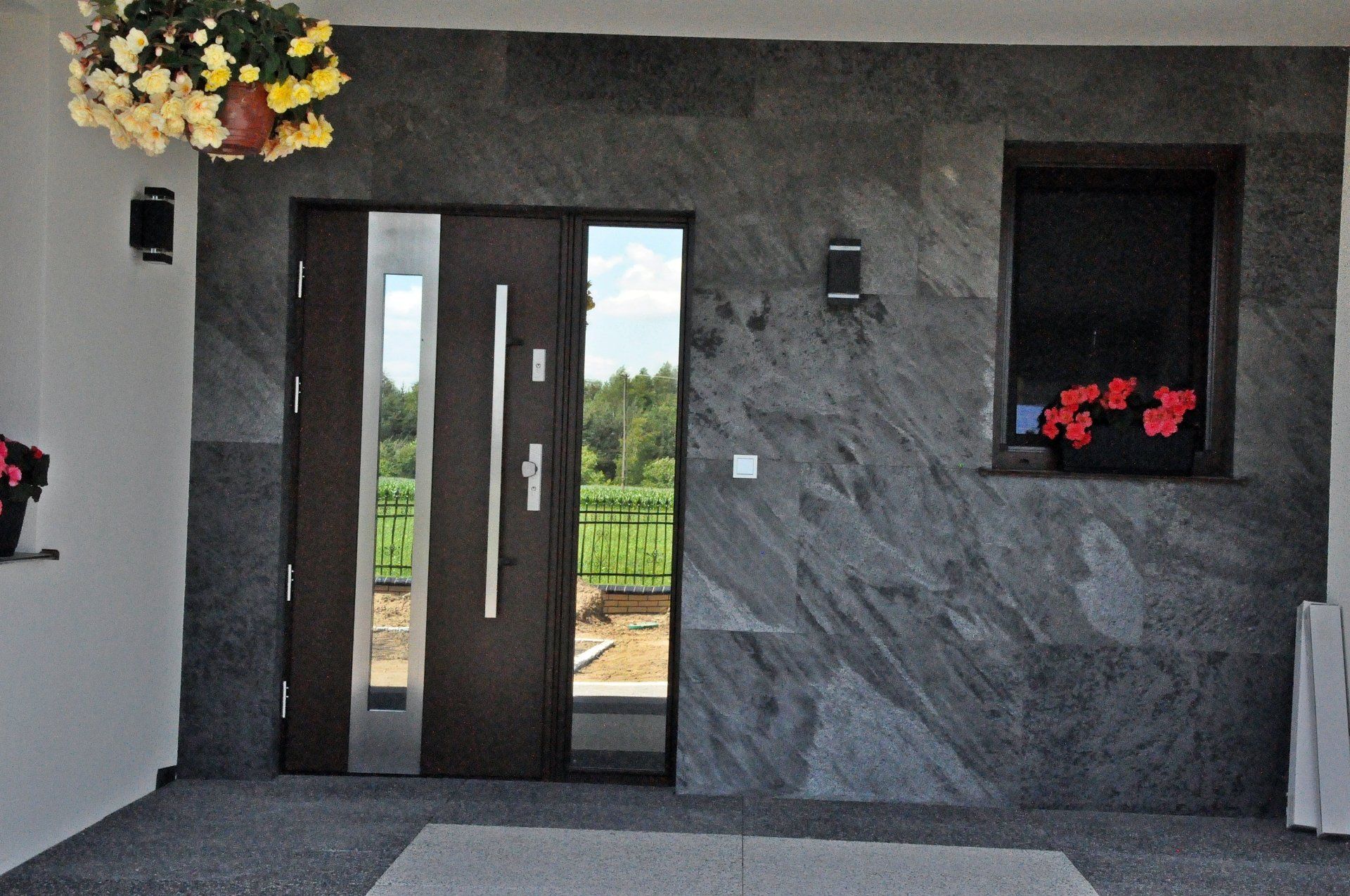 Dark-brown front door with silver accents and window beside a small window with red flowers and gray stone siding.