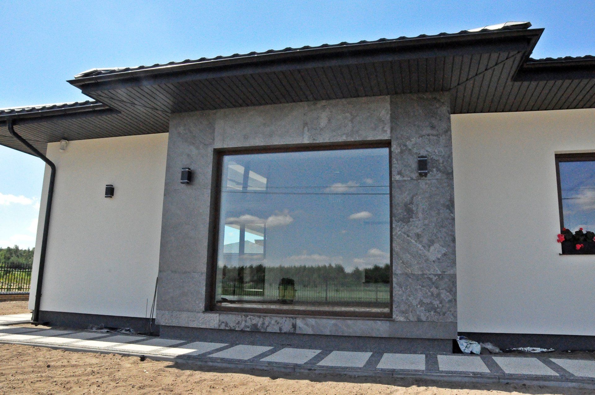 Modern house exterior with large window framed in gray stone. White walls, dark roof, and blue sky.