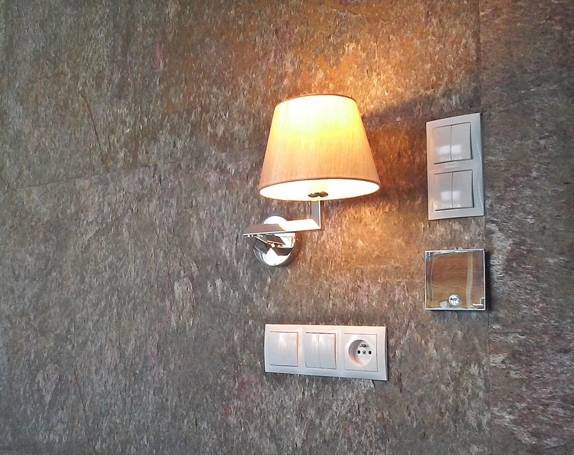 Wall-mounted lamp with beige shade above white light switches and outlet on textured, brown wall.