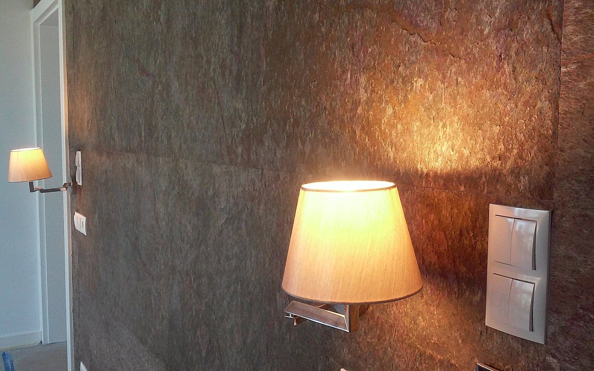 Wall with textured brown surface, lit by two sconces with tan lampshades, and light switches.