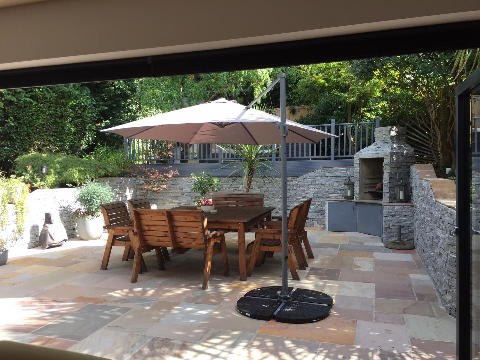 Patio with wooden table, chairs, umbrella, and stone grill, overlooking a garden.