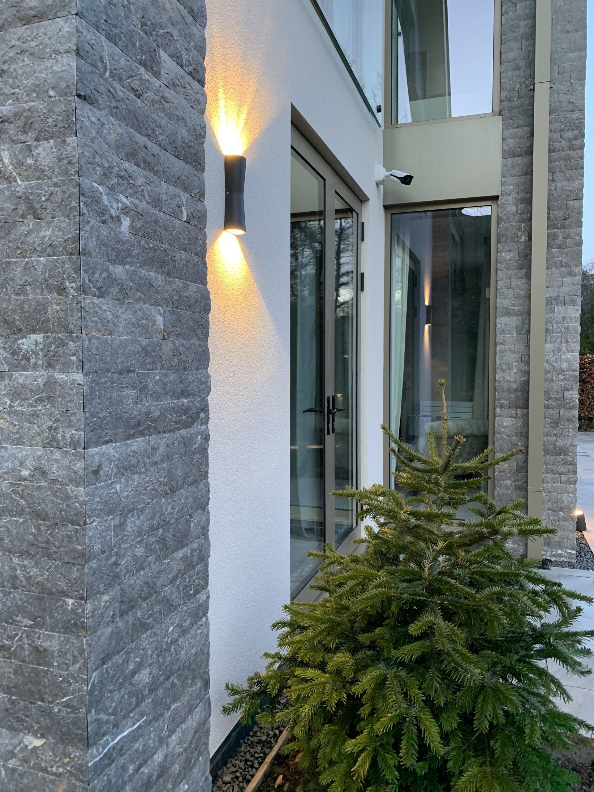 Exterior of a modern building with stone and white walls, a tree, and a light fixture.