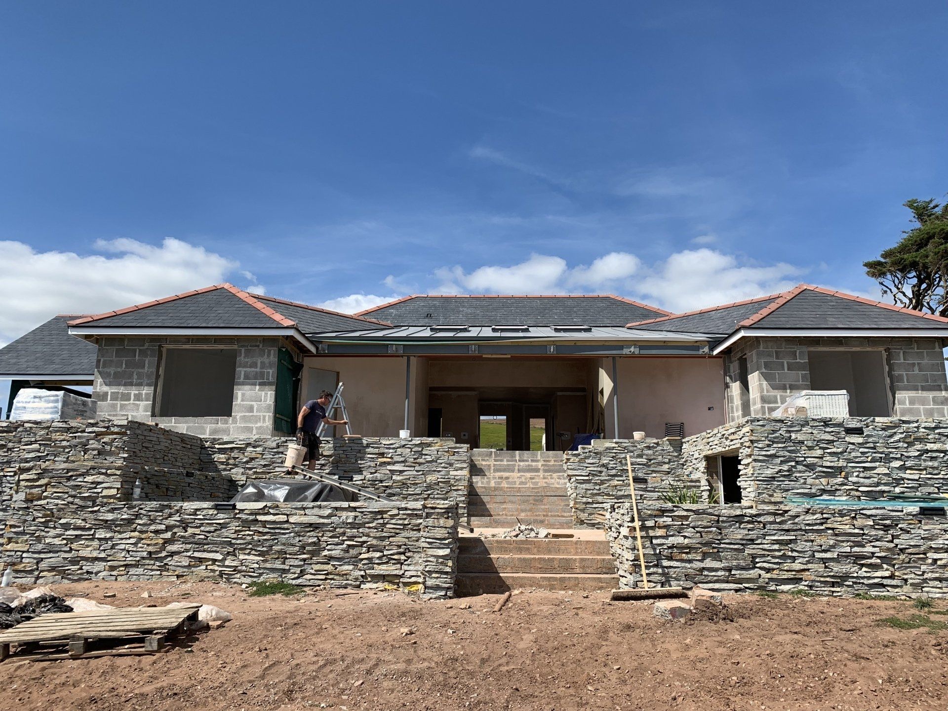Stone and block house under construction with a person working on the facade, blue sky.