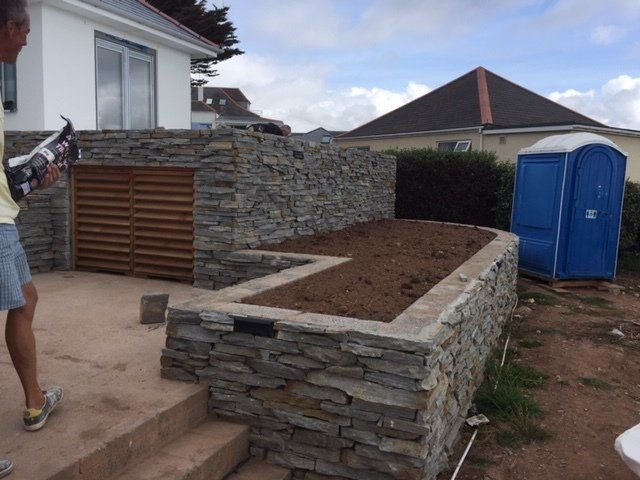 Stone retaining walls with soil bed and wooden gate, blue portable toilet, and person applying caulk.