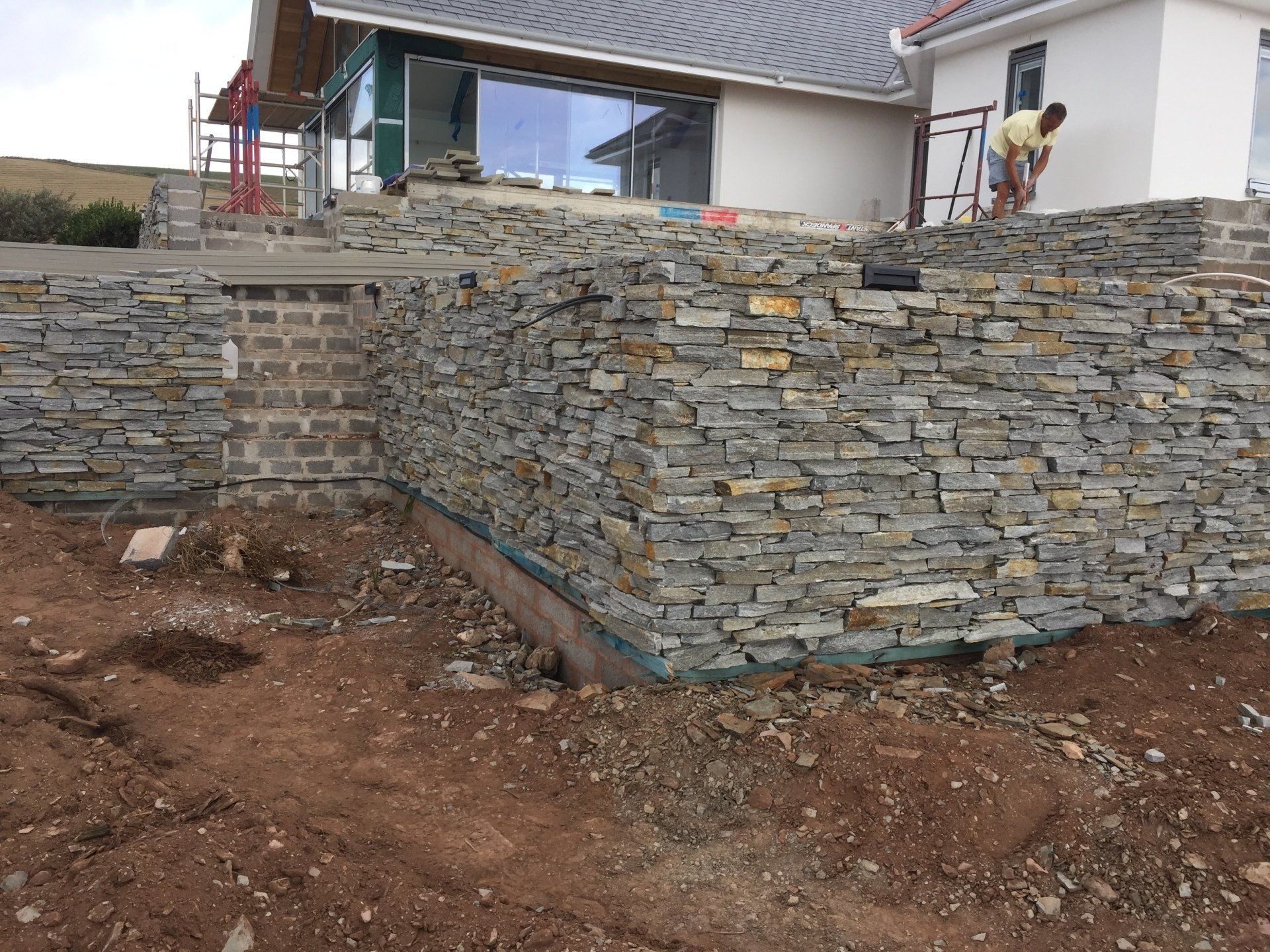 Stone retaining wall under construction with a person working near a house.