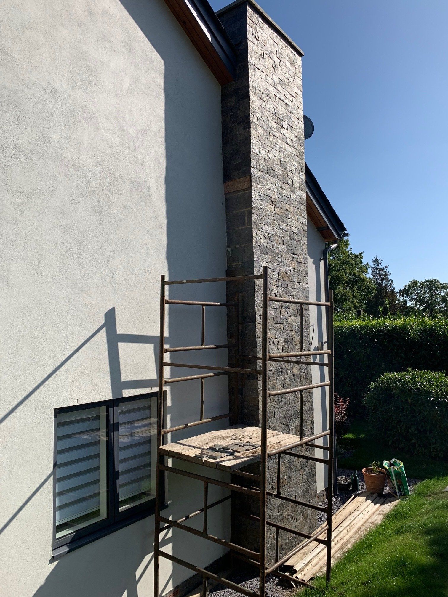 Chimney with dark stone cladding, scaffolding, next to a white stucco house, outdoors.