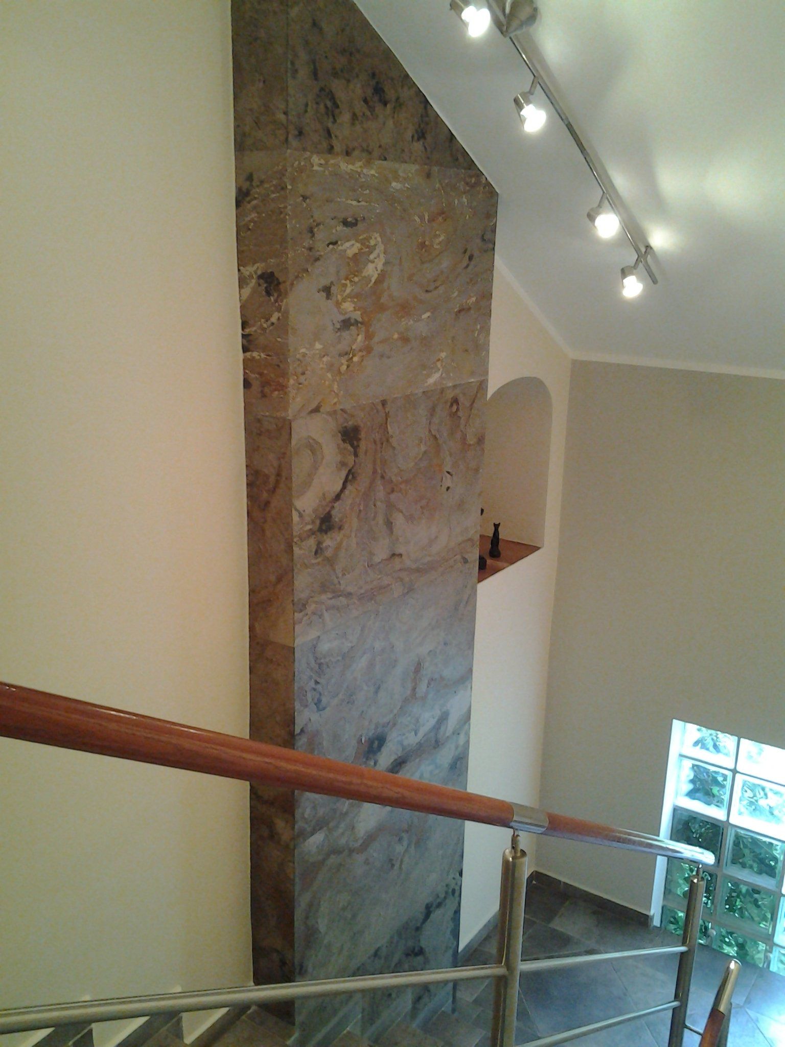 Stone-clad pillar in a stairwell, with a wooden handrail, track lighting, and a small arched niche.