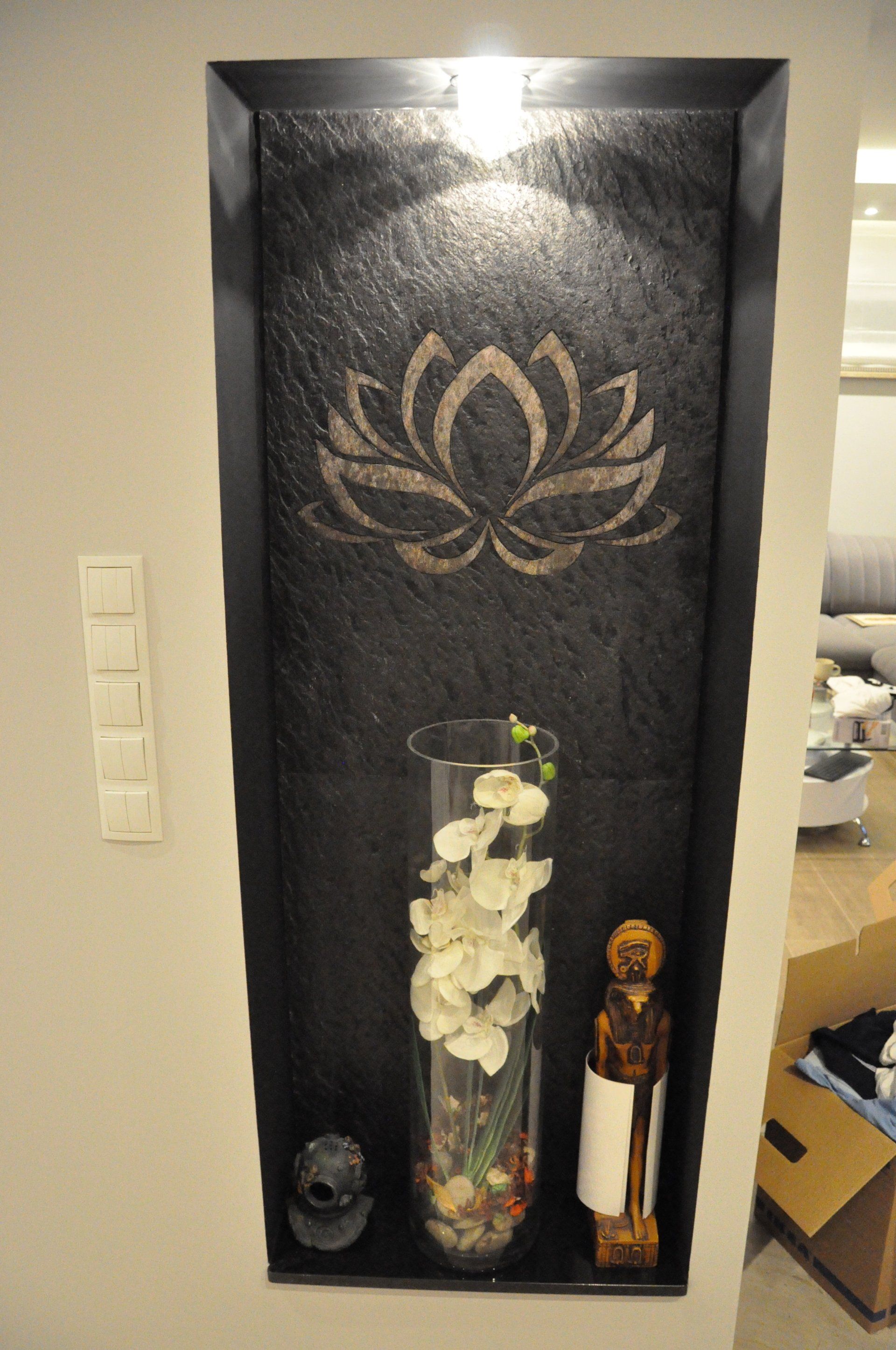 Niche in wall with black stone and lotus flower, holding vase with flowers, small statue, and light.